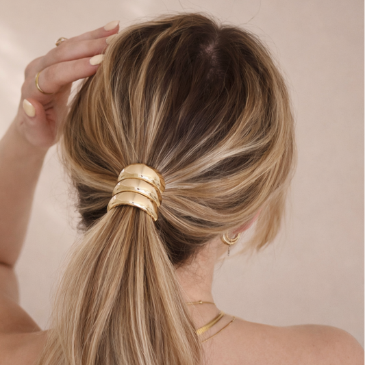 Woman with a high ponytail wearing gold hair ties and accessories against a neutral background